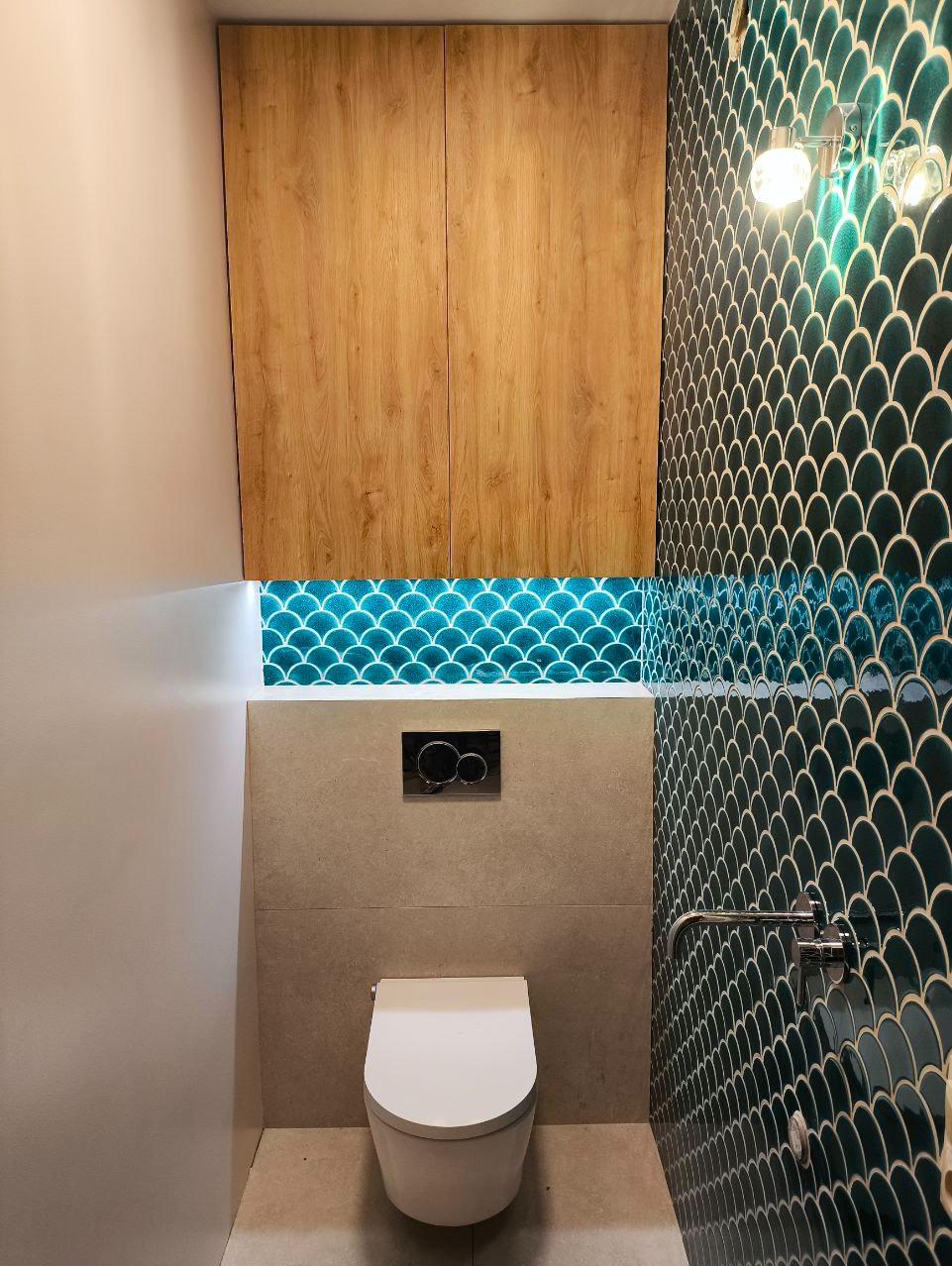 Zmodernizowana łazienka z toaletą wiszącą, szafkami z jasnego drewna nad zabudową oraz ścianą wyłożoną błyszczącymi, ciemnozielonymi płytkami w kształcie rybiej łuski, podświetlonymi od dołu.
