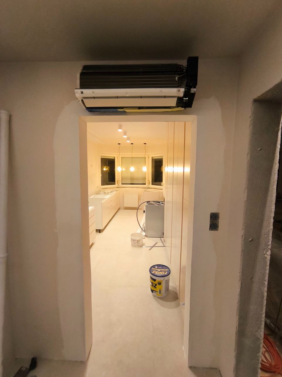 Wnętrze w trakcie prac wykończeniowych: widok przez przejście z surowymi ścianami, w tle jasna kuchnia z białymi meblami i wiszącymi lampami, na podłodze wiadro z masą szpachlową i klimatyzator...