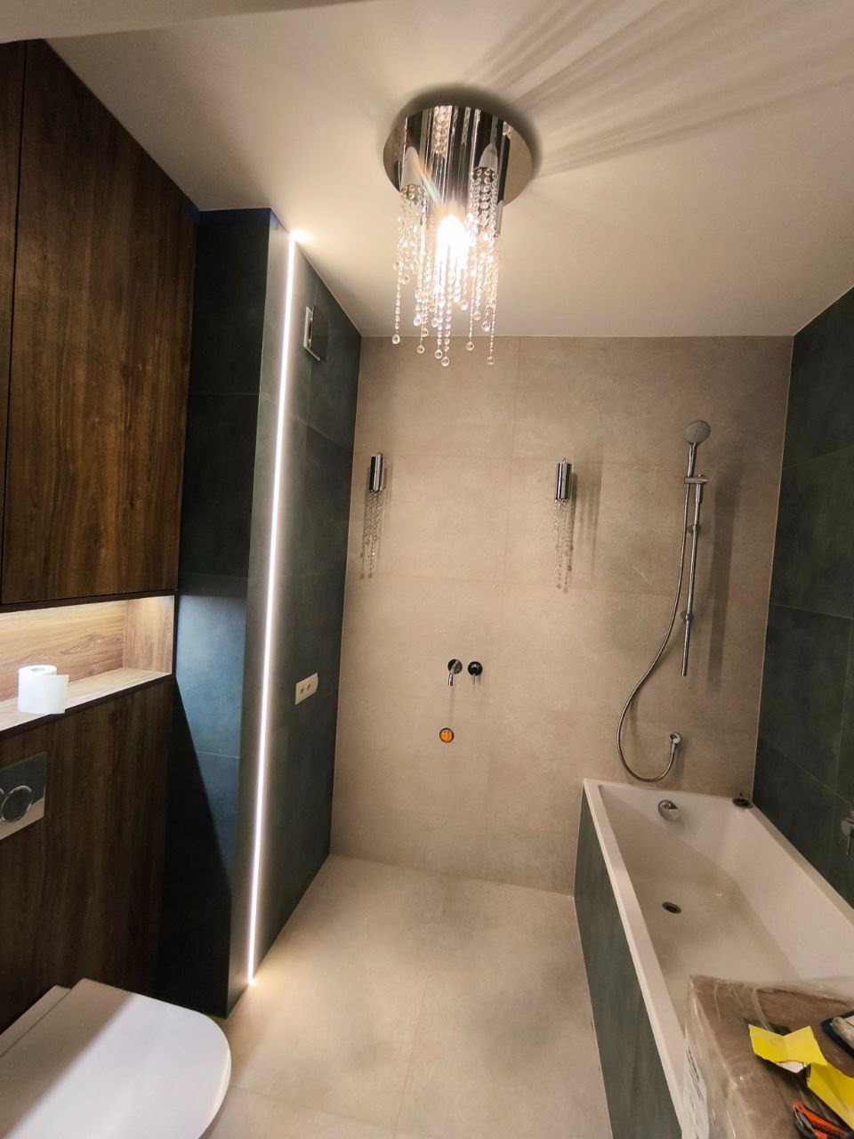 Nowoczesna łazienka z kabiną prysznicową, wanną, podwieszaną toaletą i oświetleniem LED w pionowej szczelinie ściennej oraz kryształowym żyrandolem sufitowym.