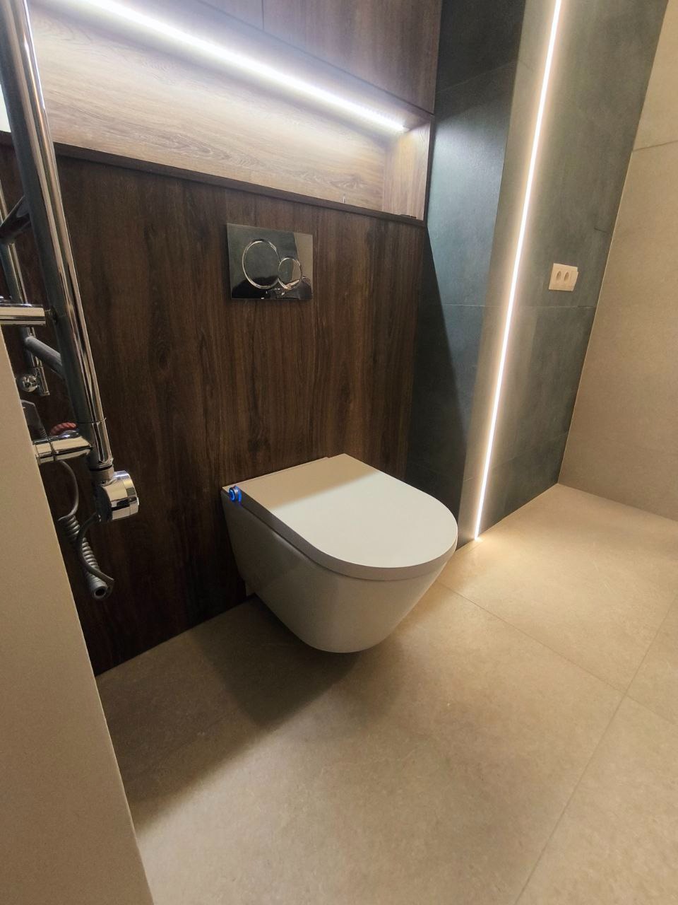 Nowoczesna toaleta podwieszana z chromowanym przyciskiem spłukiwania na tle drewnianej boazerii, oświetlona listwami LED w łazience wykończonej beżowymi płytkami.