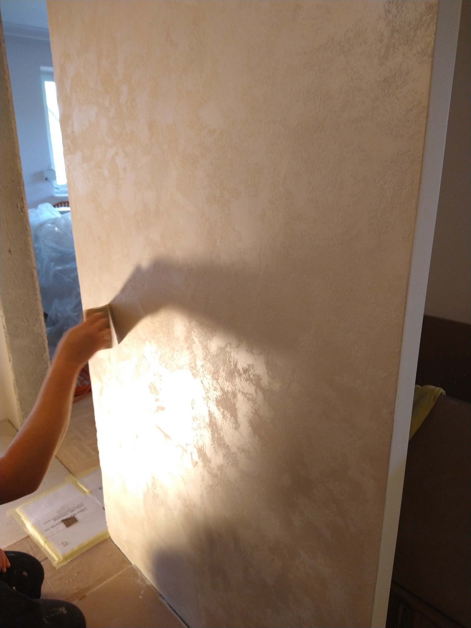 Nakładanie dekoracyjnej, beżowo-złotej powłoki na ścianę za pomocą gąbki, widoczne załamania światła, częściowo zakryte meble w tle.