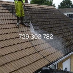 Roman Horun - Pracownik w kombinezonie i gumowych butach czyści dach z brudu za pomocą myjki ciśnieniowej w Koninie, widoczna wyraźna różnica w kolorze między czystą a brudną częścią dachu.