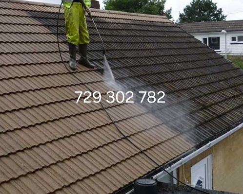 Pracownik w kombinezonie i gumowych butach czyści dach z brudu za pomocą myjki ciśnieniowej w Koninie, widoczna wyraźna różnica w kolorze między czystą a brudną częścią dachu.