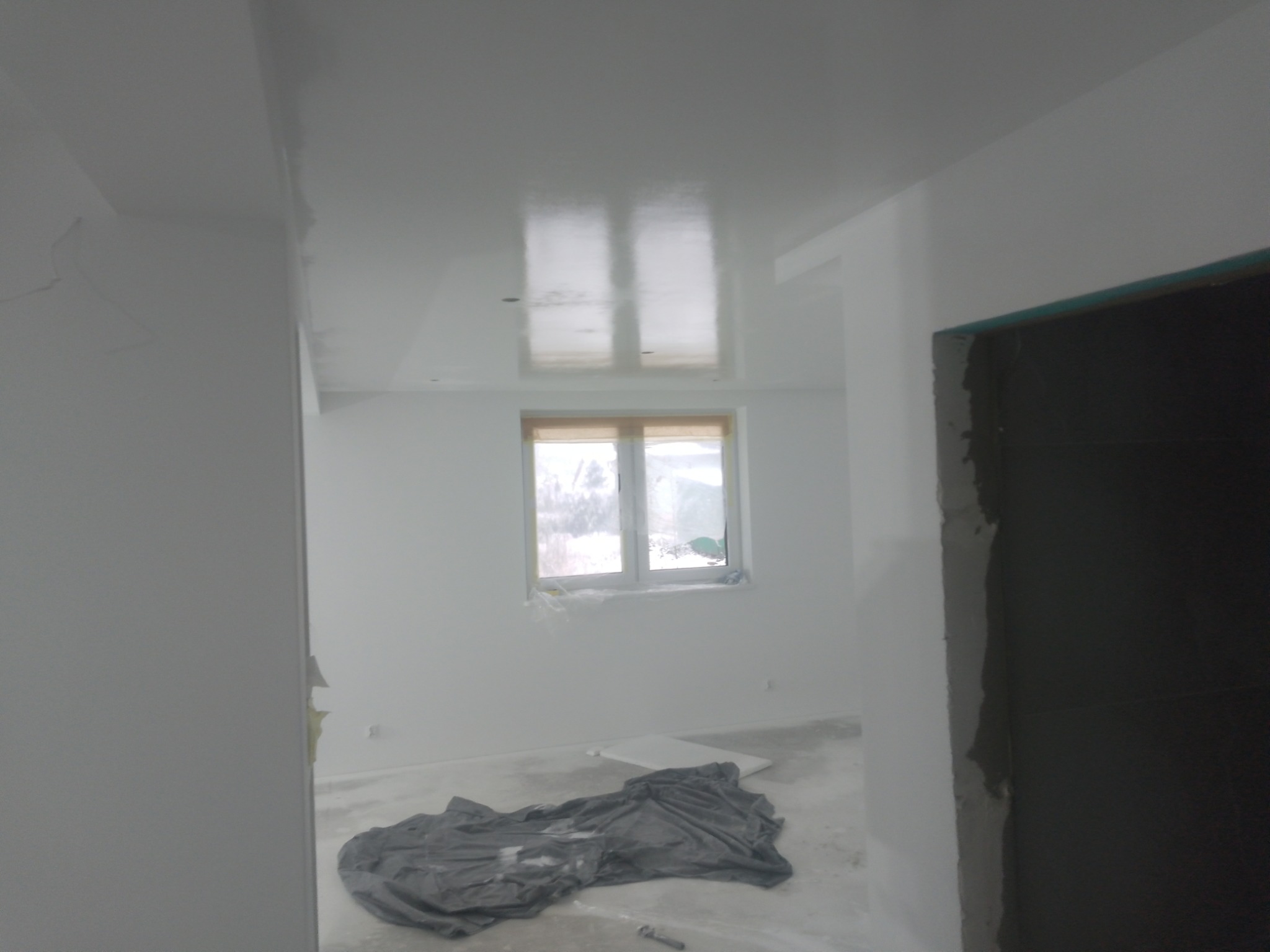 Wnętrze pokoju w trakcie malowania na biało, widoczna folia ochronna na podłodze, świeżo pomalowany sufit odbija światło z okna.