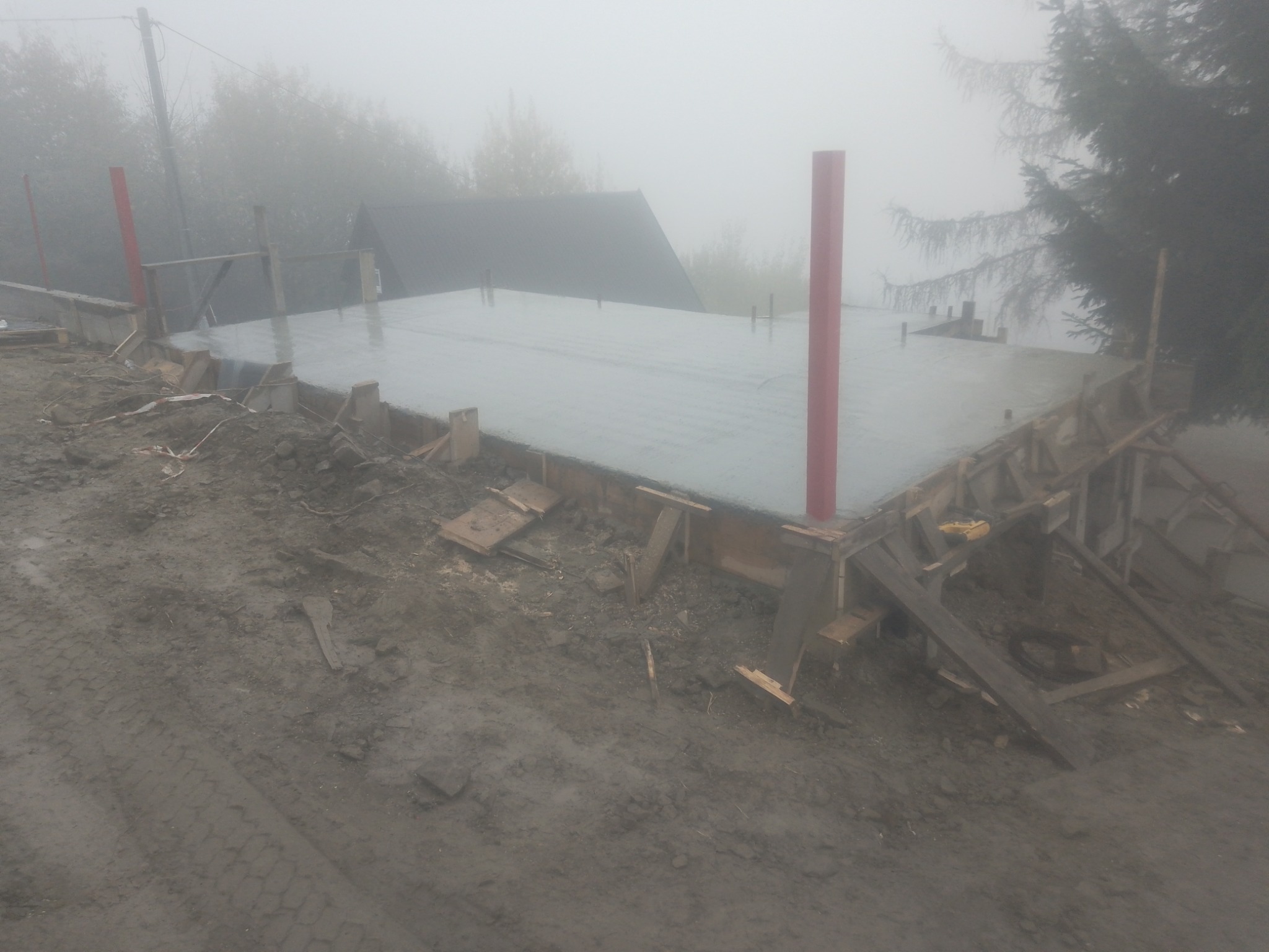 Świeżo wylana, mokra płyta fundamentowa z czerwonymi słupkami w narożach, otoczona drewnianym szalunkiem i błotnistym terenem w gęstej mgle.