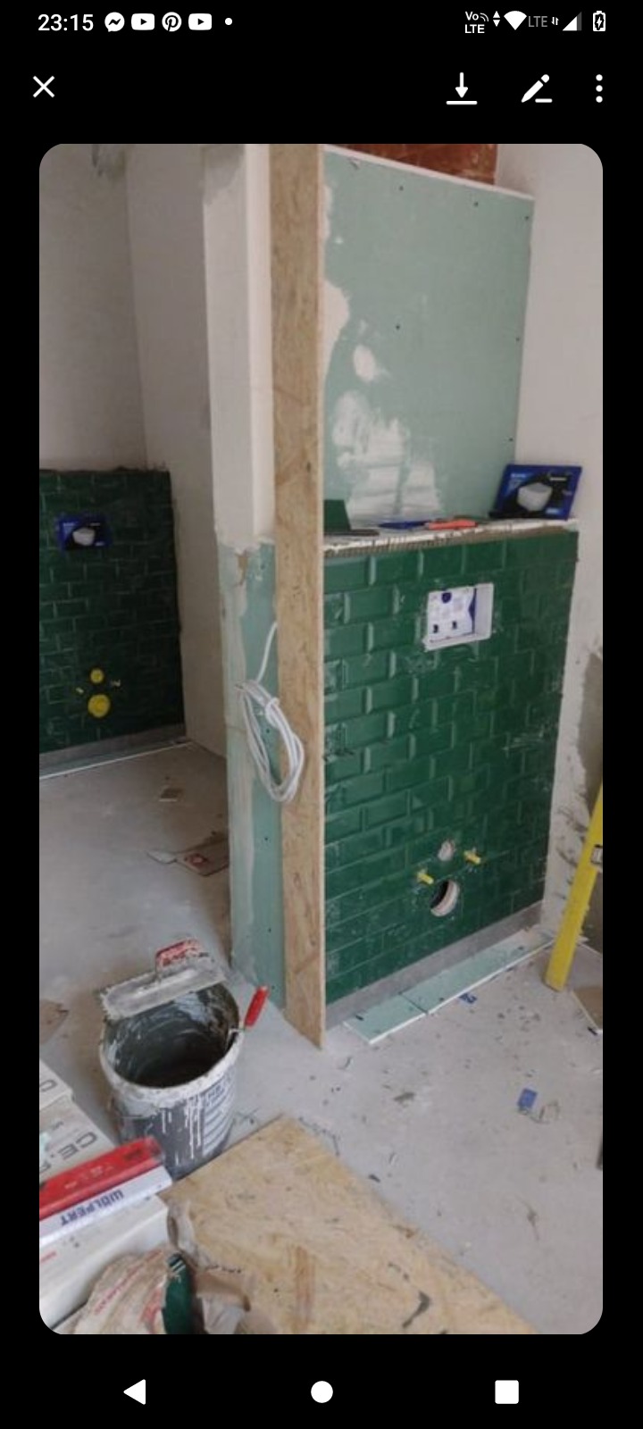 Wnętrze łazienki w trakcie remontu: ściany wyłożone ciemnozielonymi, błyszczącymi płytkami typu metro, widoczne instalacje podtynkowe, płyta gipsowo-kartonowa i narzędzia budowlane.