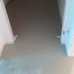 Antoni Przybylski - Wylewka samopoziomująca w pomieszczeniu, widoczne rury instalacyjne przy ścianie, surowy stan wykończenia, przygotowanie do dalszych prac remontowych.