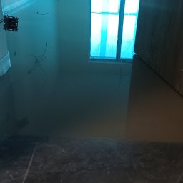 Antoni Przybylski - Wylana, samopoziomująca wylewka podłogowa w pomieszczeniu z zabezpieczonymi oknami folią. Widoczne kable elektryczne i fragment wykończonej podłogi.