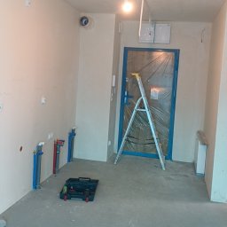 Antoni Przybylski - Wnętrze w trakcie remontu: ściany przygotowane do malowania, widoczne rury instalacji wodnej, drabina oparta o zabezpieczone folią drzwi, skrzynka z narzędziami na podłodze.