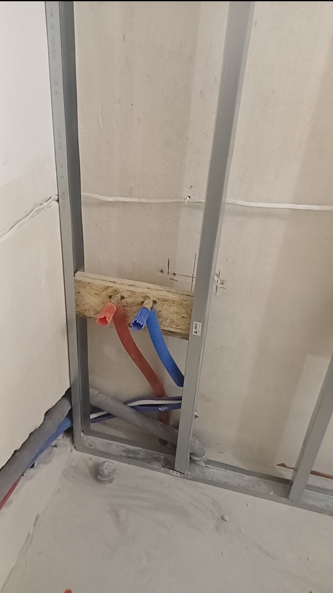 Konstrukcja metalowa z rurami wodnymi (czerwona i niebieska) w ścianie. Widoczne elementy instalacji wodnej w trakcie prac remontowych. Surowy stan, przygotowanie do dalszych prac.