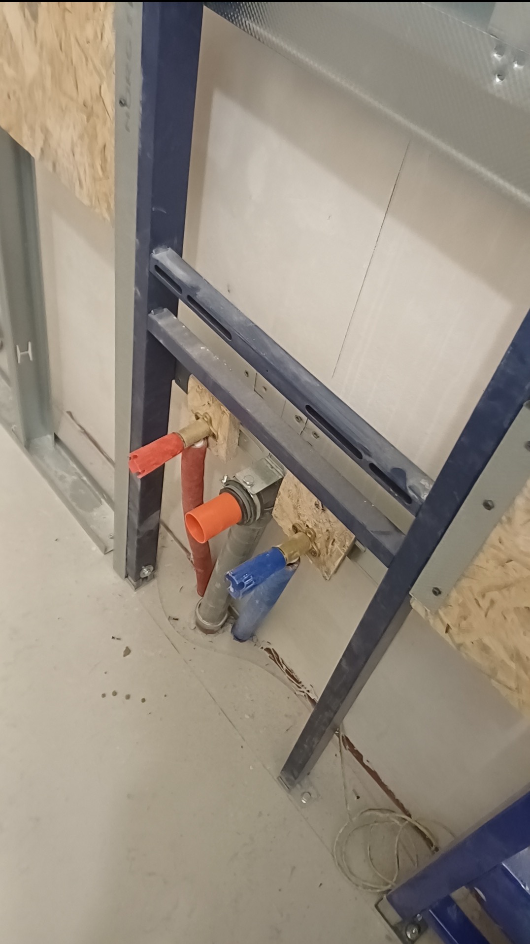 Stelaż podtynkowy z instalacją wodną (czerwona, pomarańczowa, niebieska rura) zamontowany przy ścianie z płyt OSB i karton-gipsu. Widok z góry na podłogę.