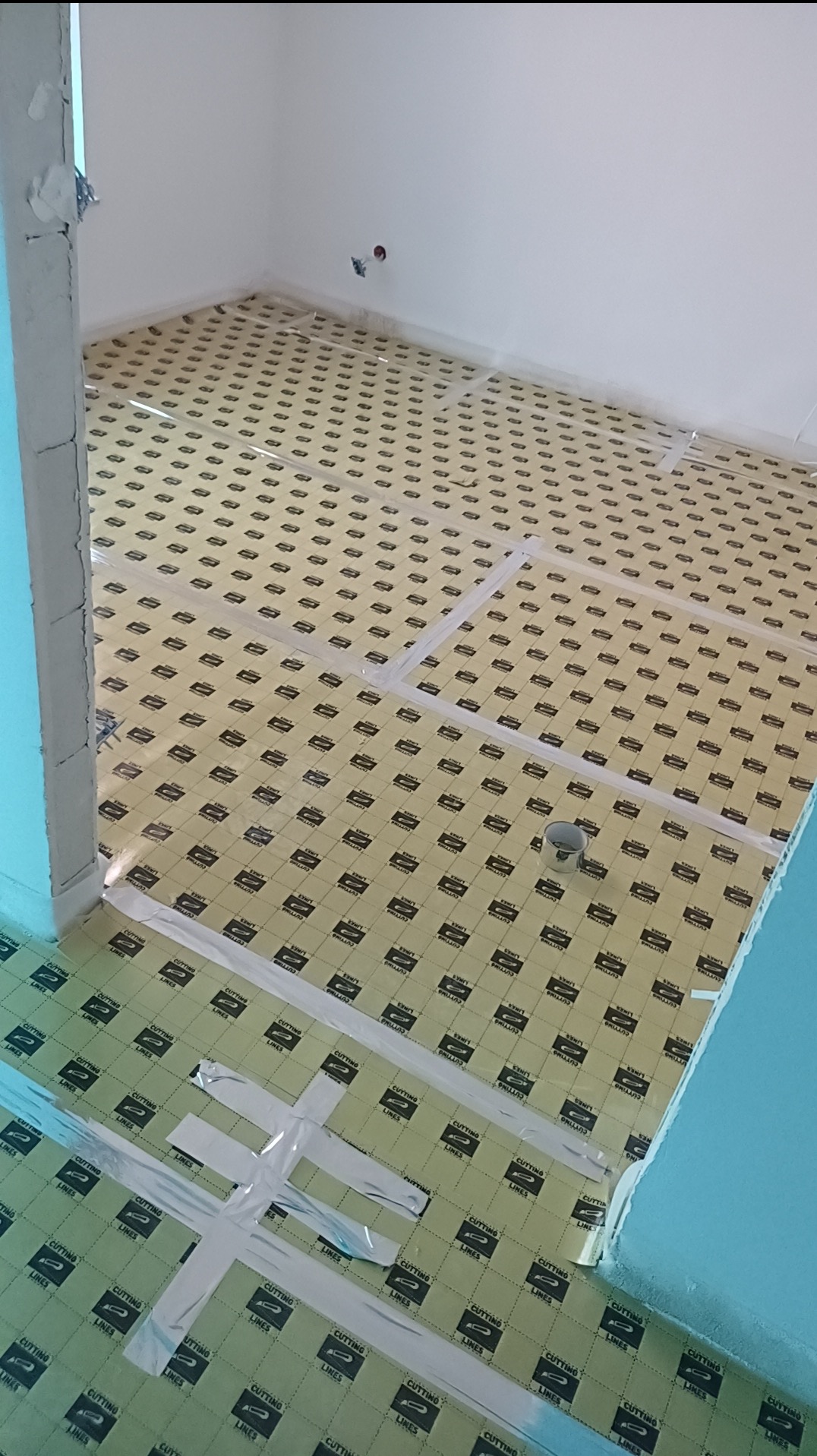 Przygotowanie podłoża pod panele: rozłożona mata izolacyjna z taśmą mocującą w pustym pokoju. Widoczne gniazdka elektryczne na ścianie i fragment puszki z farbą.