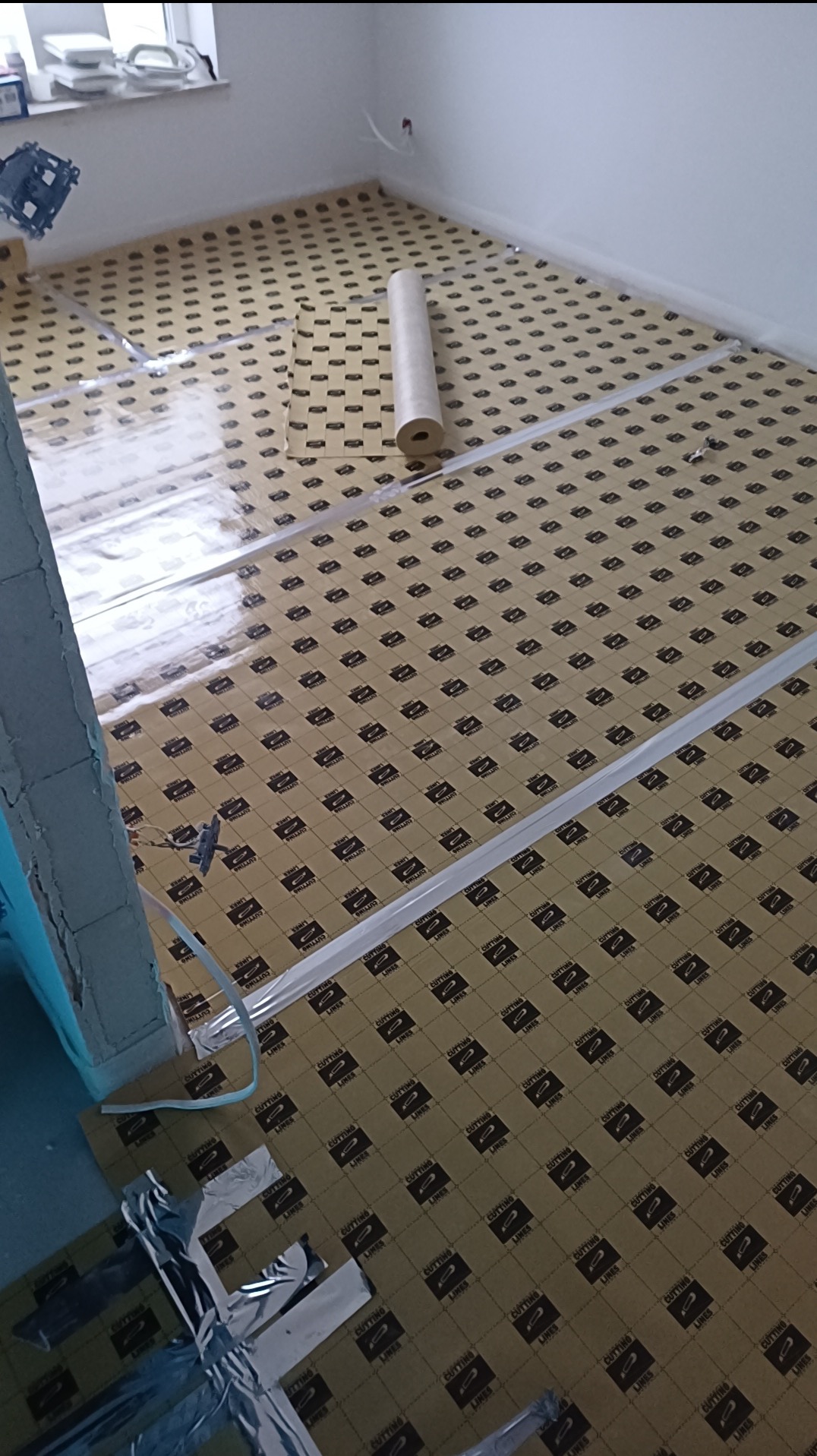 Przygotowanie podłoża pod panele: rozłożona mata wygłuszająca z folią paroizolacyjną, widoczne taśmy klejące. Etap prac przed ułożeniem paneli podłogowych.