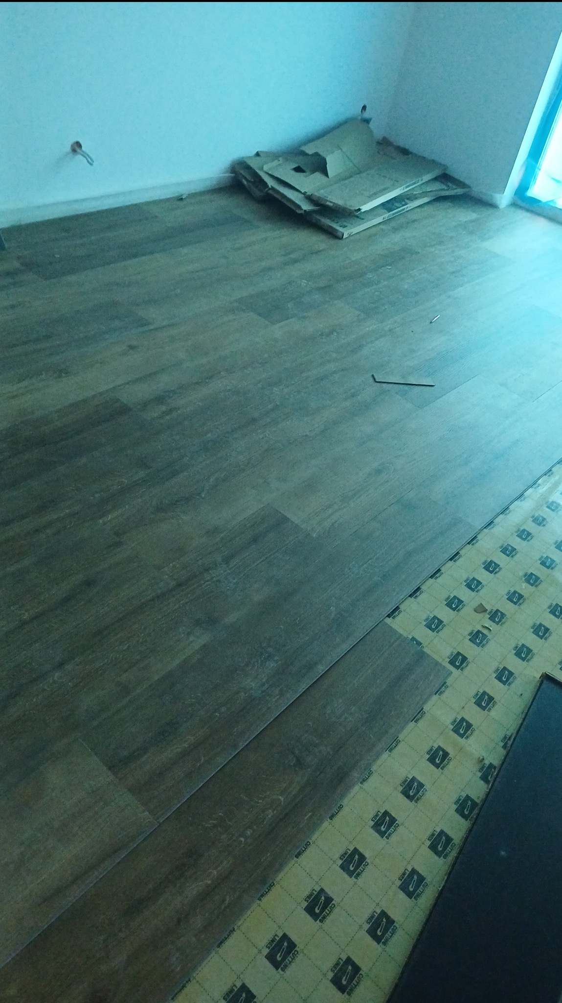 Układanie paneli podłogowych w trakcie. Część podłogi wyłożona panelami w odcieniach brązu, widoczny podkład i panele do ułożenia. W rogu sterta kartonów.