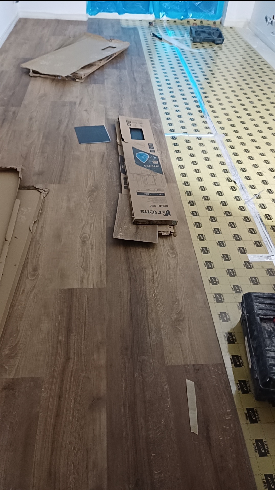 Układanie paneli podłogowych: widok z góry na brązowe panele, podkład, kartony po panelach i narzędzia. Częściowo ułożona podłoga z widocznym podkładem.