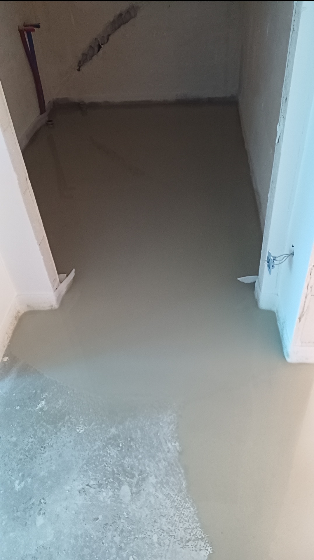 Wylewka samopoziomująca w pomieszczeniu, widoczne rury instalacyjne przy ścianie, surowy stan wykończenia, przygotowanie do dalszych prac remontowych.