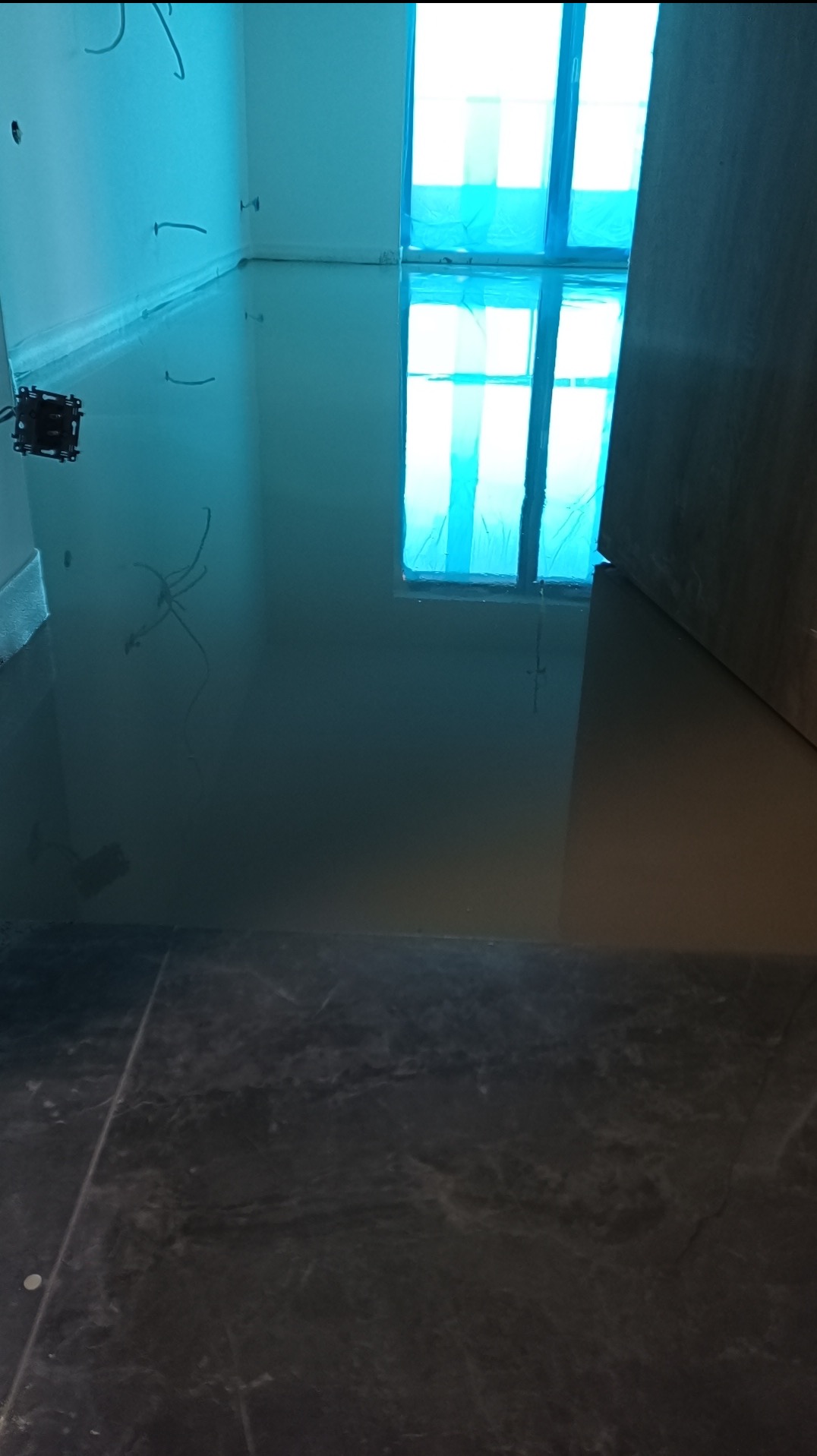 Wylana, samopoziomująca wylewka podłogowa w pomieszczeniu z zabezpieczonymi oknami folią. Widoczne kable elektryczne i fragment wykończonej podłogi.