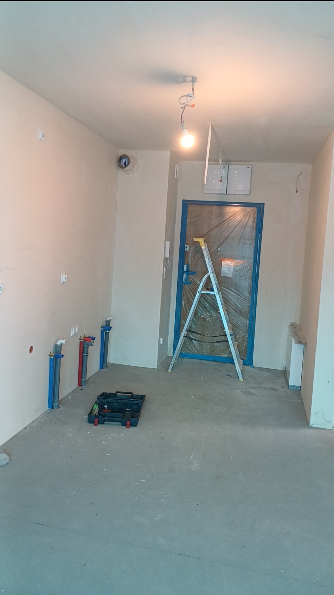 Wnętrze w trakcie remontu: ściany przygotowane do malowania, widoczne rury instalacji wodnej, drabina oparta o zabezpieczone folią drzwi, skrzynka z narzędziami na podłodze.