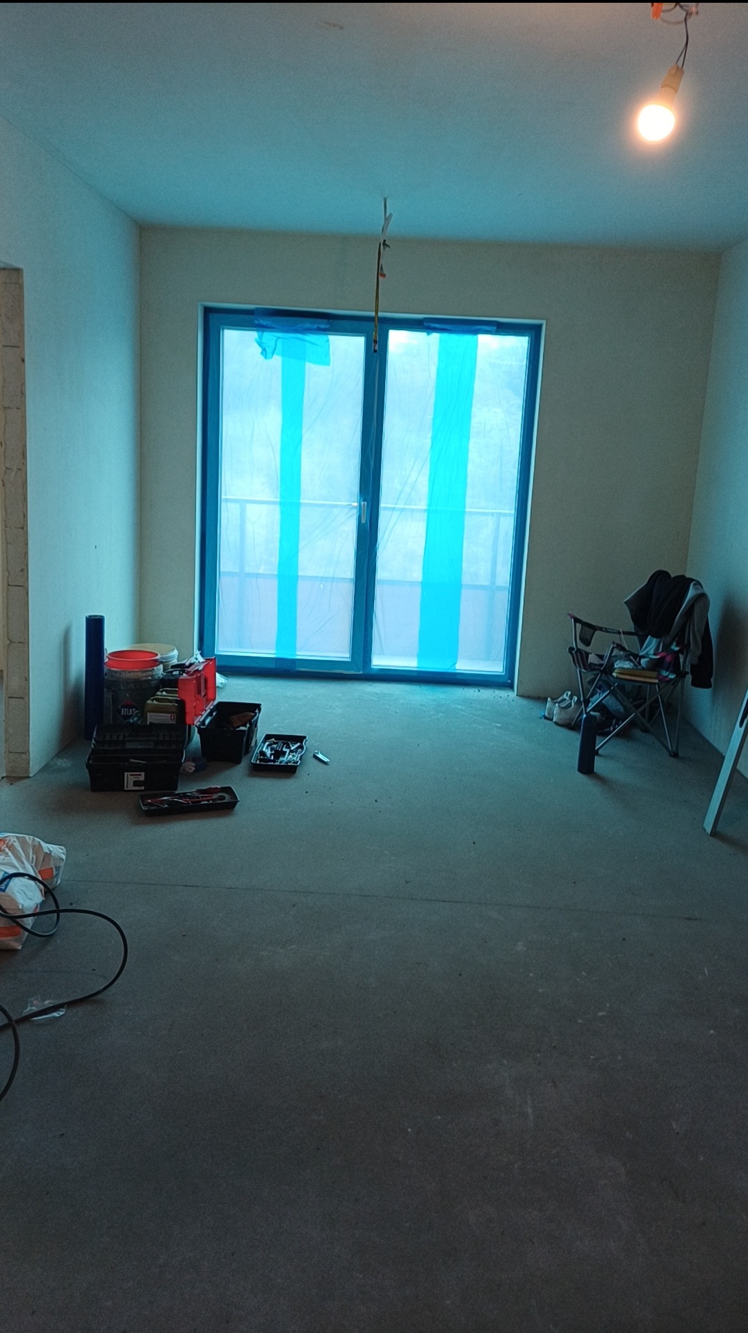 Pokój w trakcie remontu: zabezpieczone okno folią, narzędzia malarskie, surowa podłoga, ściany przygotowane do malowania, prowizoryczne oświetlenie.