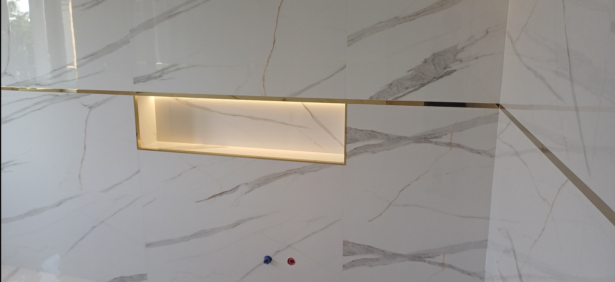 Biała ściana z marmurowym wzorem, złote listwy dekoracyjne i podświetlana wnęka. Widoczne fragmenty instalacji wodnej.