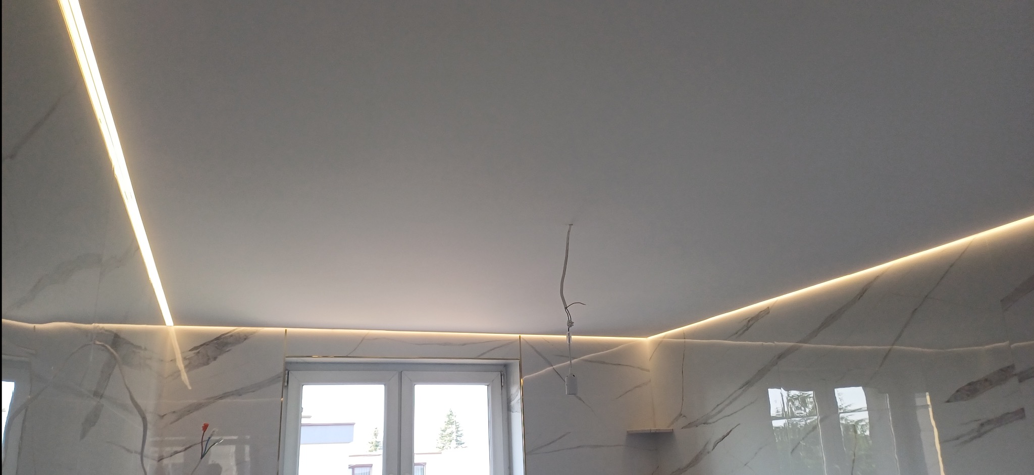 Nowoczesne oświetlenie LED wzdłuż sufitu i ściany wykończonej płytkami imitującymi marmur. Widoczne okno i wystający kabel od oświetlenia.