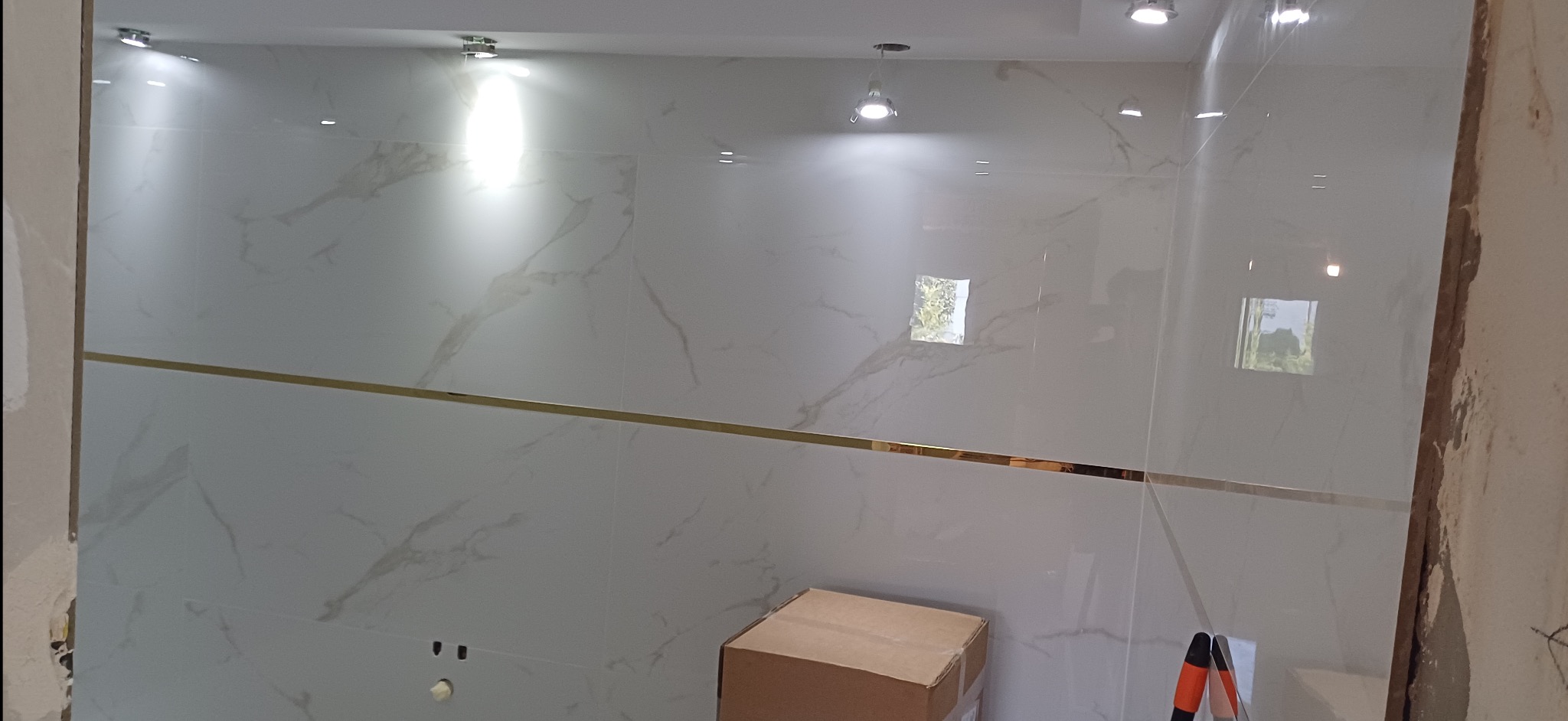 Wykończona ściana łazienki z białych płytek imitujących marmur, z poziomym złotym paskiem dekoracyjnym i zamontowanym oświetleniem punktowym. Widoczny karton i fragment drabiny.