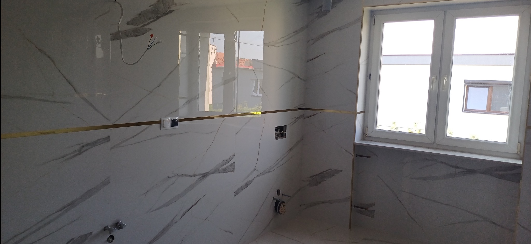 Wykończona łazienka z białymi płytkami imitującymi marmur, złotymi akcentami i oknem. Widoczne instalacje pod armaturę. Nowoczesny design.