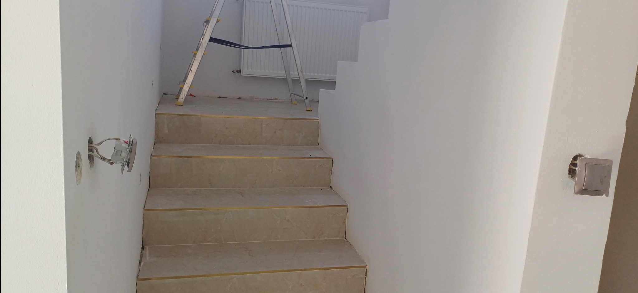 Klatka schodowa w trakcie remontu: wyłożone płytki na schodach z metalowymi listwami, ściany po szpachlowaniu i malowaniu, widoczne puszki elektryczne, drabina.
