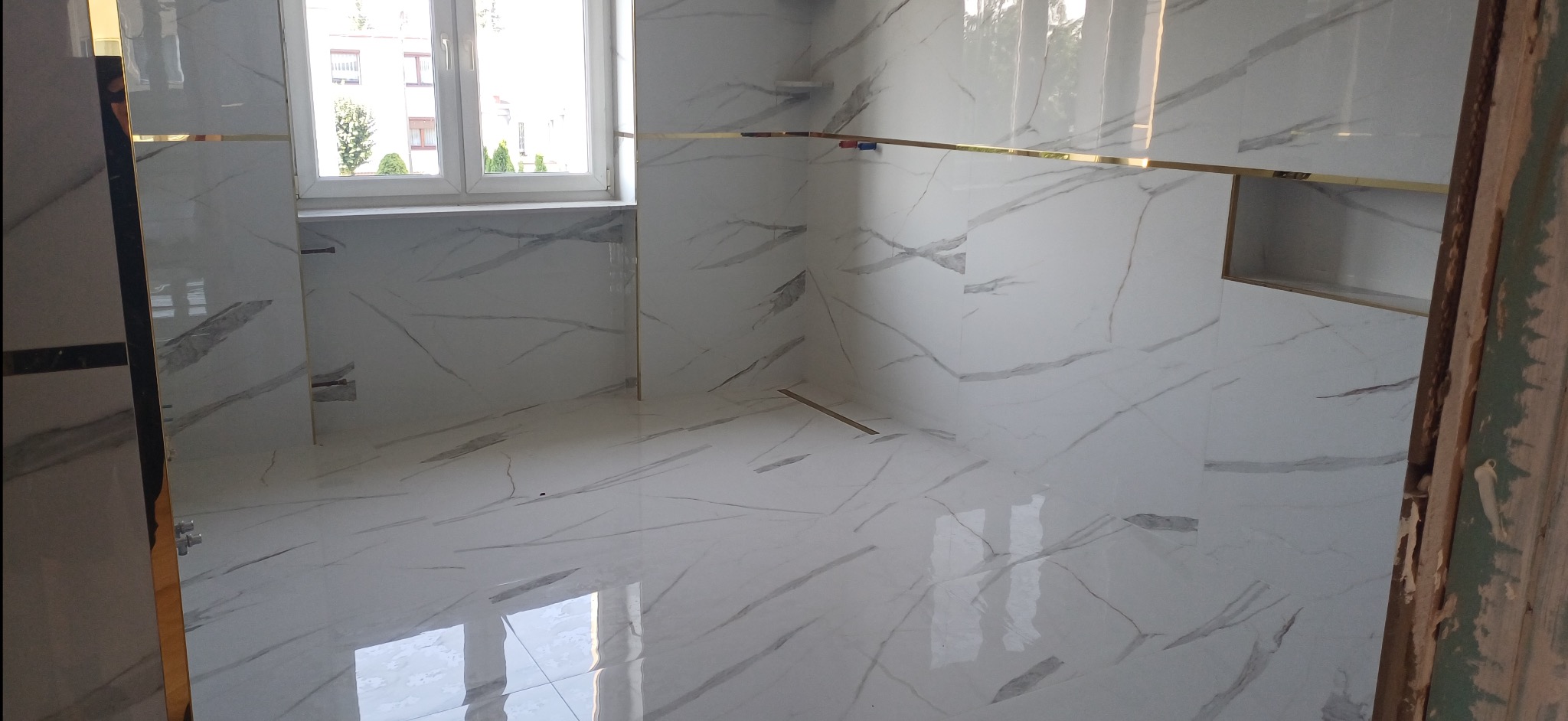 Wykończona łazienka z białymi płytkami imitującymi marmur, złotymi listwami i wnęką. Widoczne okno z widokiem na zieleń. Ujęcie z perspektywy wejścia, fragment ościeżnicy.