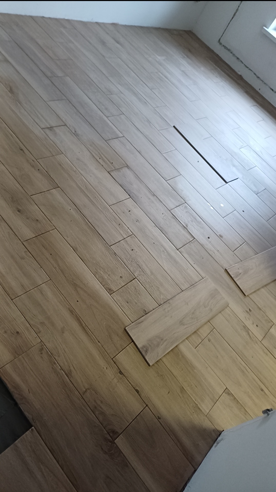 Układanie paneli podłogowych w jodełkę, widoczne niedokończone fragmenty przy ścianie i otwór wentylacyjny. Jasne panele imitujące drewno.