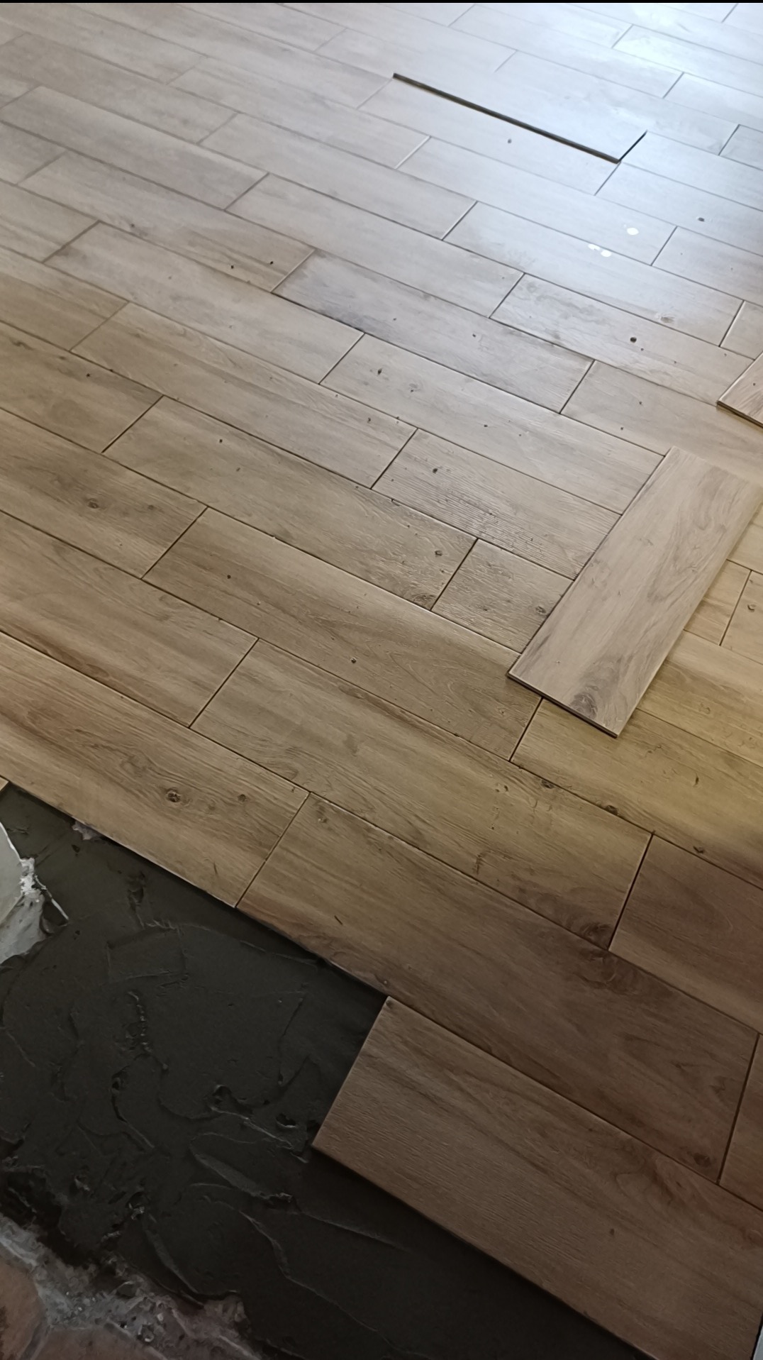 Układanie podłogi z płytek imitujących drewno. Widoczny klej i fragmenty ułożonej podłogi. Płytki ułożone w jodełkę, ciepłe odcienie brązu.