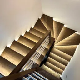 Drewniane schody z nowoczesnym oświetleniem LED pod każdym stopniem, widok z góry, białe ściany, eleganckie wykończenie. Unikalny design oświetlenia schodów.