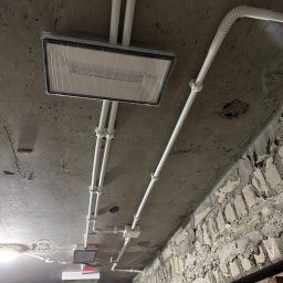IVElectric Sp. z o.o. - Montaż lampy LED z instalacją w białych rurkach na surowym, betonowym suficie. Widoczne kable i mocowania. Surowy stan wykończenia ścian.
