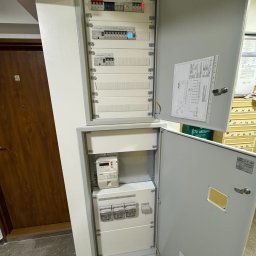 IVElectric Sp. z o.o. - Otwarta skrzynka z bezpiecznikami i licznikiem energii elektrycznej w korytarzu, widoczne schematy i dokumentacja. Szara obudowa, białe wnętrze, brązowe drzwi w tle.