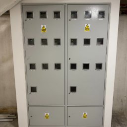 IVElectric Sp. z o.o. - Szara szafa elektryczna z wieloma licznikami oznaczonymi numerami lokali, umieszczona w rogu pomieszczenia. Widoczne ostrzeżenia o napięciu elektrycznym.