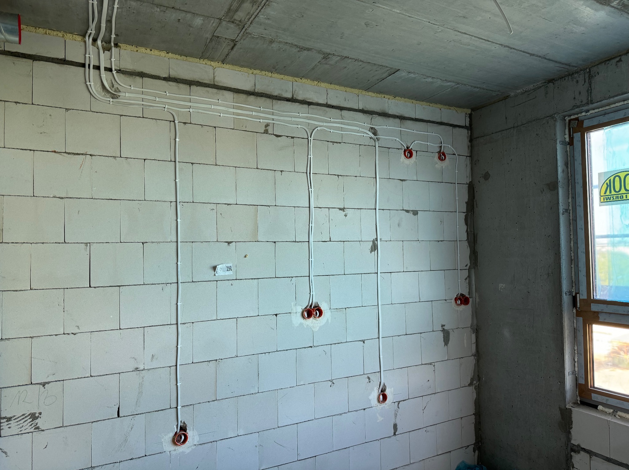 Surowy stan: Białe kable elektryczne poprowadzone po ścianie z pustaków, czerwone puszki pod gniazdka, widok na okno w surowym stanie.