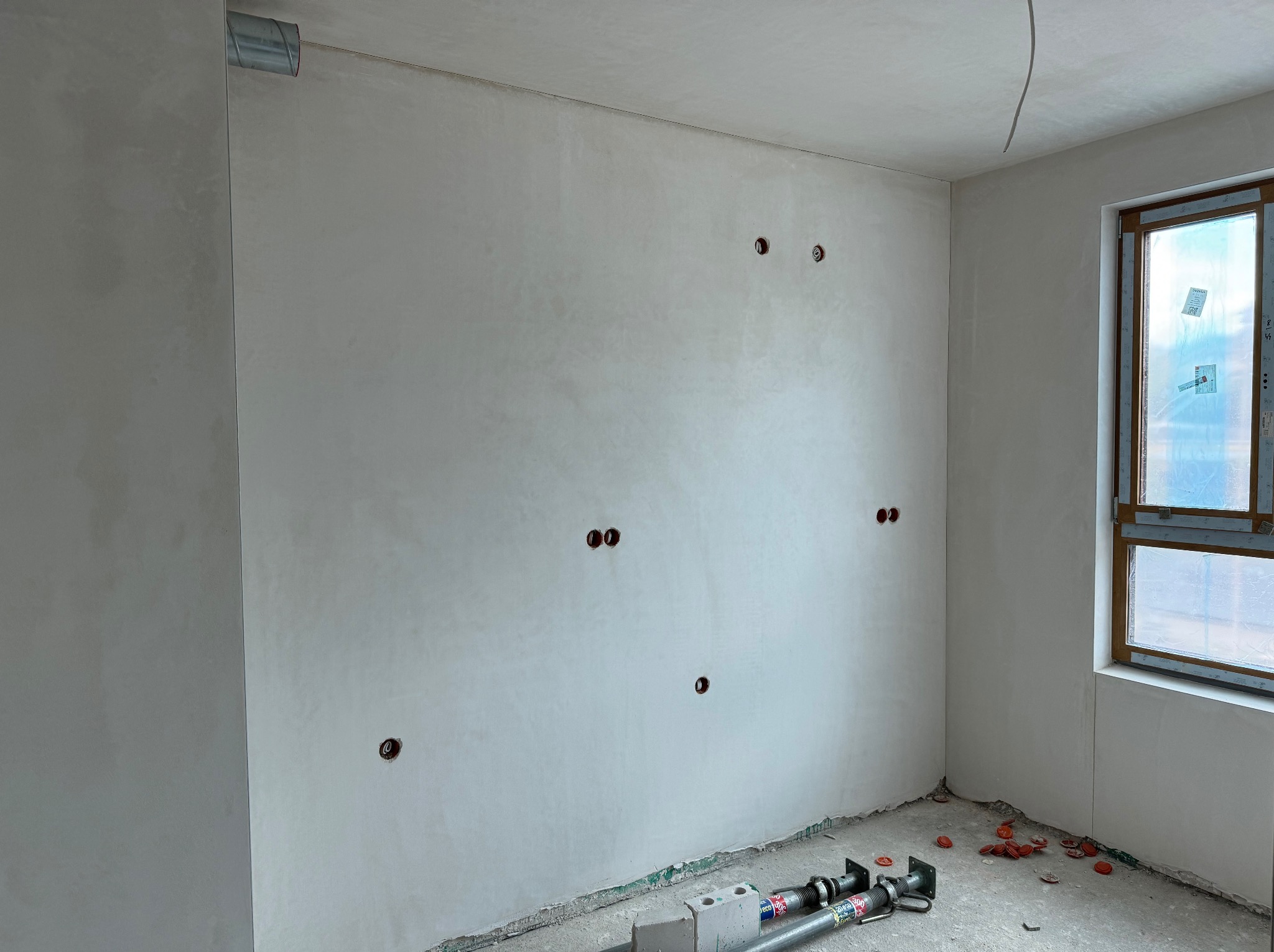 Surowy stan wnętrza z widocznymi otworami pod gniazdka elektryczne w świeżo otynkowanej ścianie, rurą wentylacyjną przy suficie i zabezpieczonym oknem, na podłodze leżą resztki po pracach i podpory...