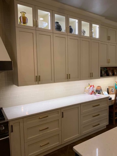 Kuchnia z kremowymi szafkami, szklanymi witrynami podświetlanymi od wewnątrz i blatem z jasnego kamienia, oświetlonym listwą LED podszafkową.