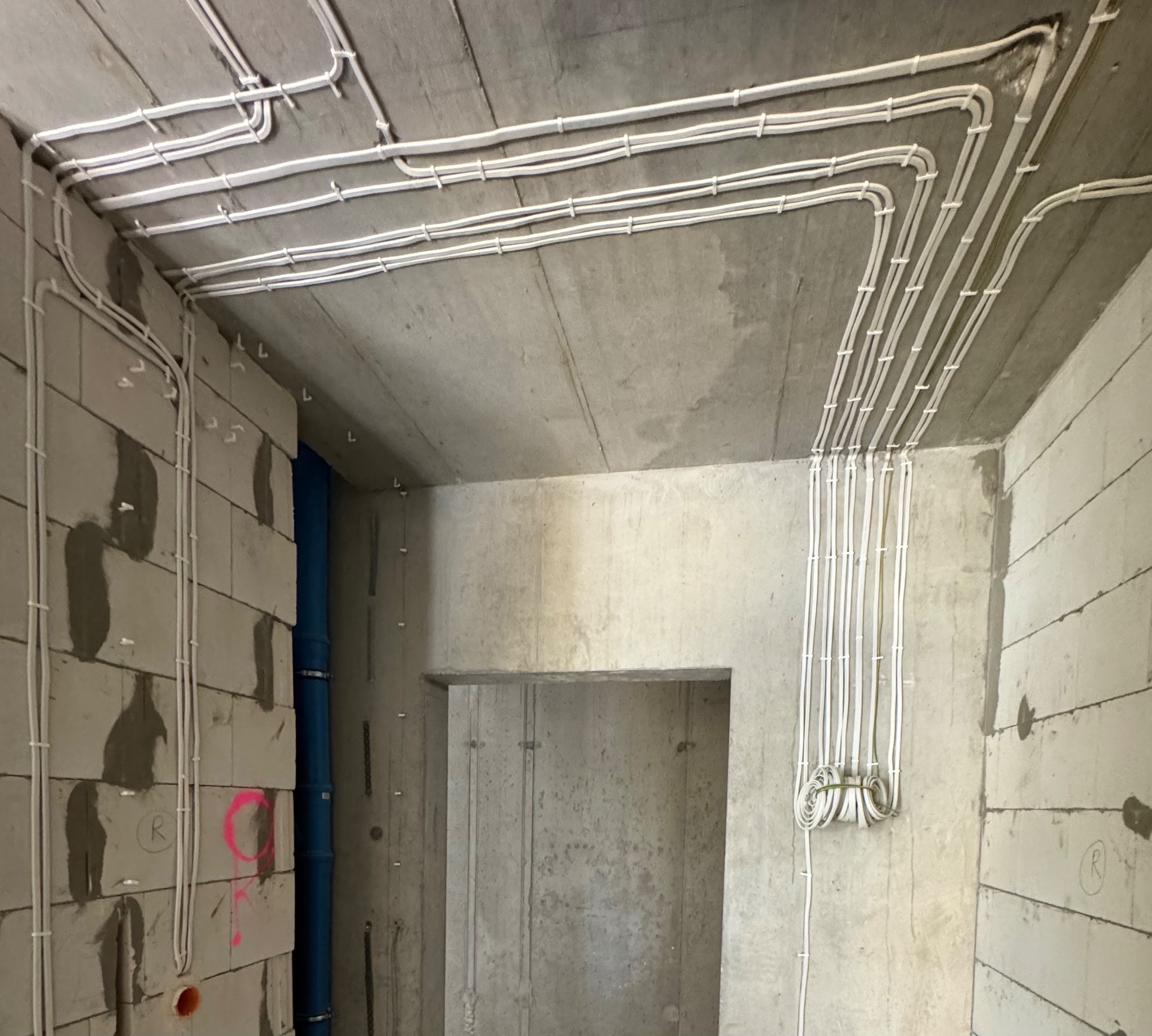 Surowe wnętrze w trakcie prac wykończeniowych z widocznymi, nowymi przewodami elektrycznymi poprowadzonymi po ścianach i suficie, przytwierdzonymi uchwytami, z nieotynkowanymi ścianami z betonu...