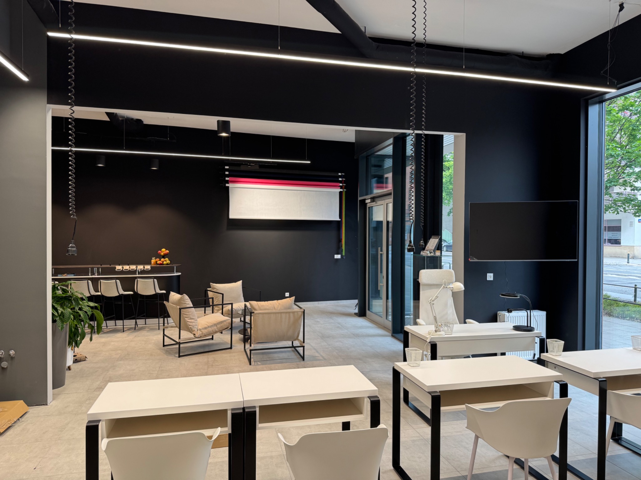 Nowoczesne biuro z czarnymi ścianami i liniowym oświetleniem LED. Widoczne białe biurka i krzesła, bar z owocami i designerskie fotele. Minimalistyczny design.