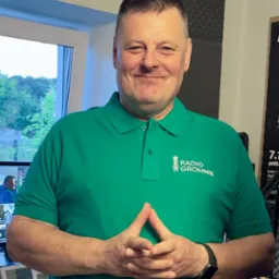 Uśmiechnięty mężczyzna w zielonej koszulce polo z logo 'Radio Gromnik', w tle okno i sprzęt radiowy.