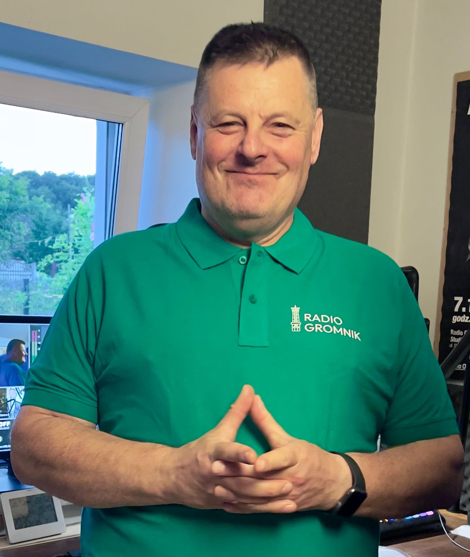 Uśmiechnięty mężczyzna w zielonej koszulce polo z logo 'Radio Gromnik', w tle okno i sprzęt radiowy.