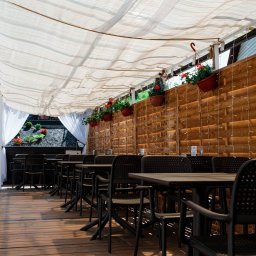 Restauracja Food Makerzy Kluszkowce - Zadaszony taras restauracji z drewnianą podłogą, czarnymi krzesłami i stołami, dekorowany kwiatami w wiszących donicach i delikatnymi lampkami.