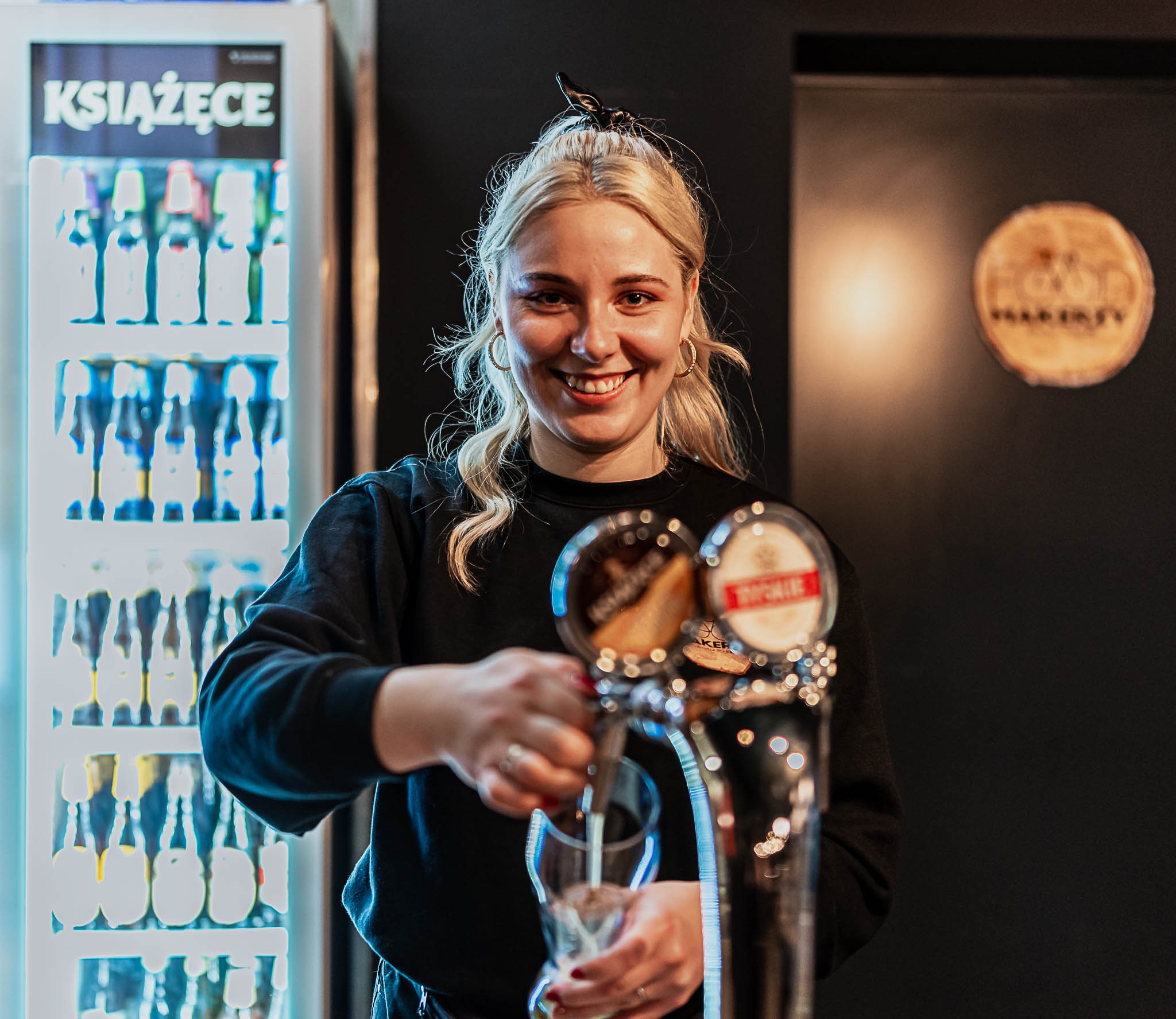 Uśmiechnięta barmanka nalewa piwo z kranu do szklanki, w tle lodówka z napojami marki Książęce i logo lokalu na czarnej ścianie.