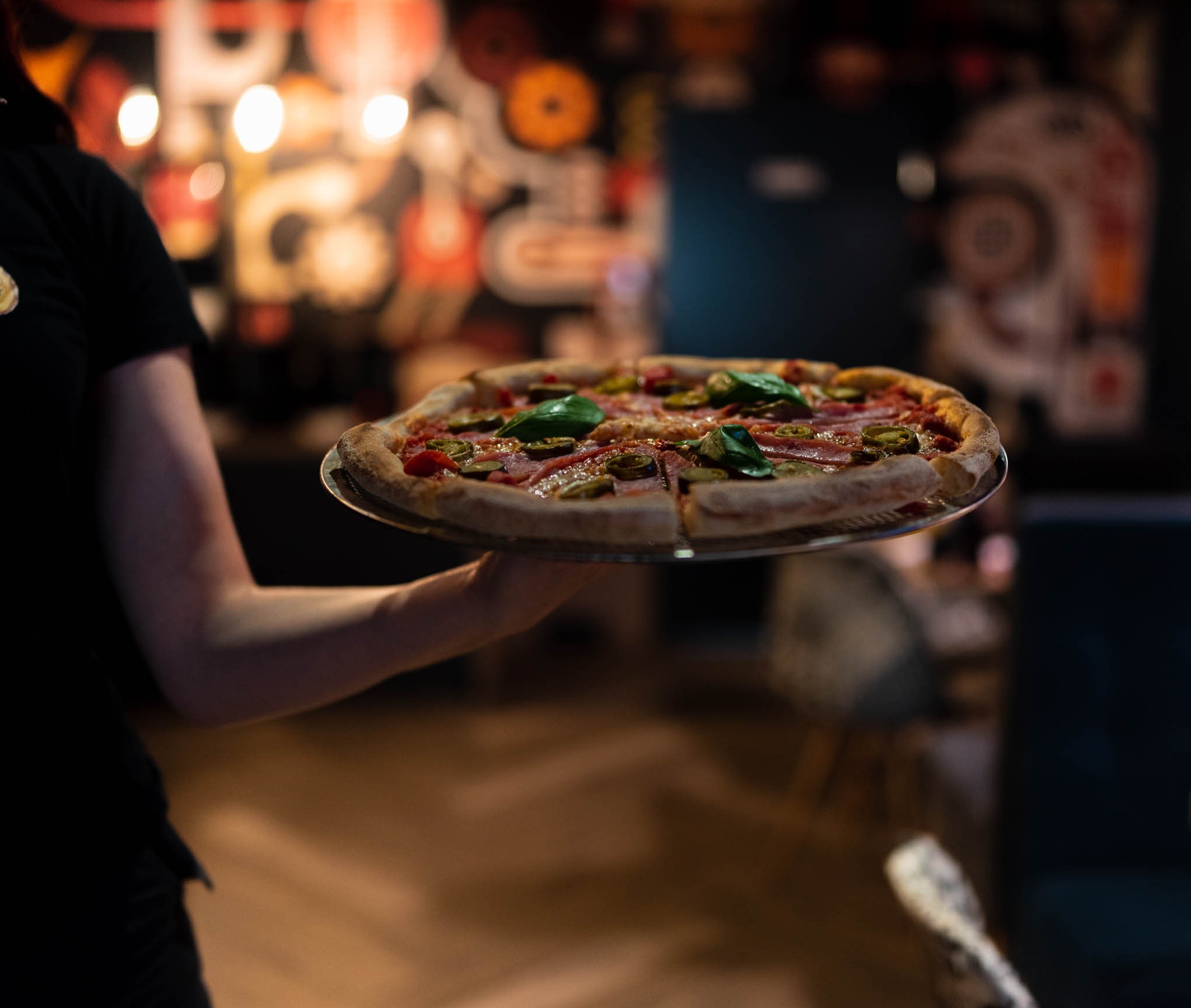 Kelnerka w czarnej koszulce niesie pizzę z szynką, papryczkami jalapeno i bazylią na metalowej tacy w restauracji z rozmytym, kolorowym tłem.