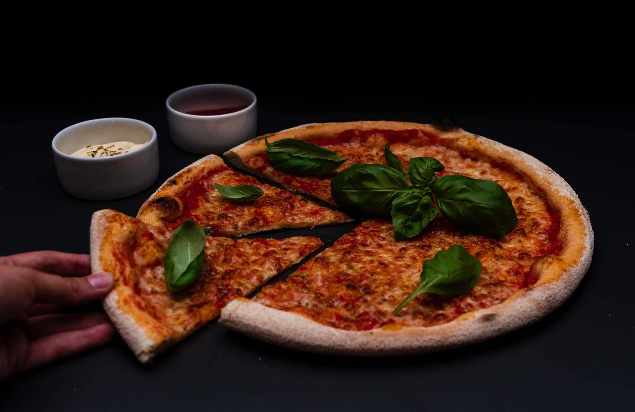 Pizza margherita z bazylią na czarnym tle, dwa sosy w białych miseczkach, dłoń sięga po kawałek.