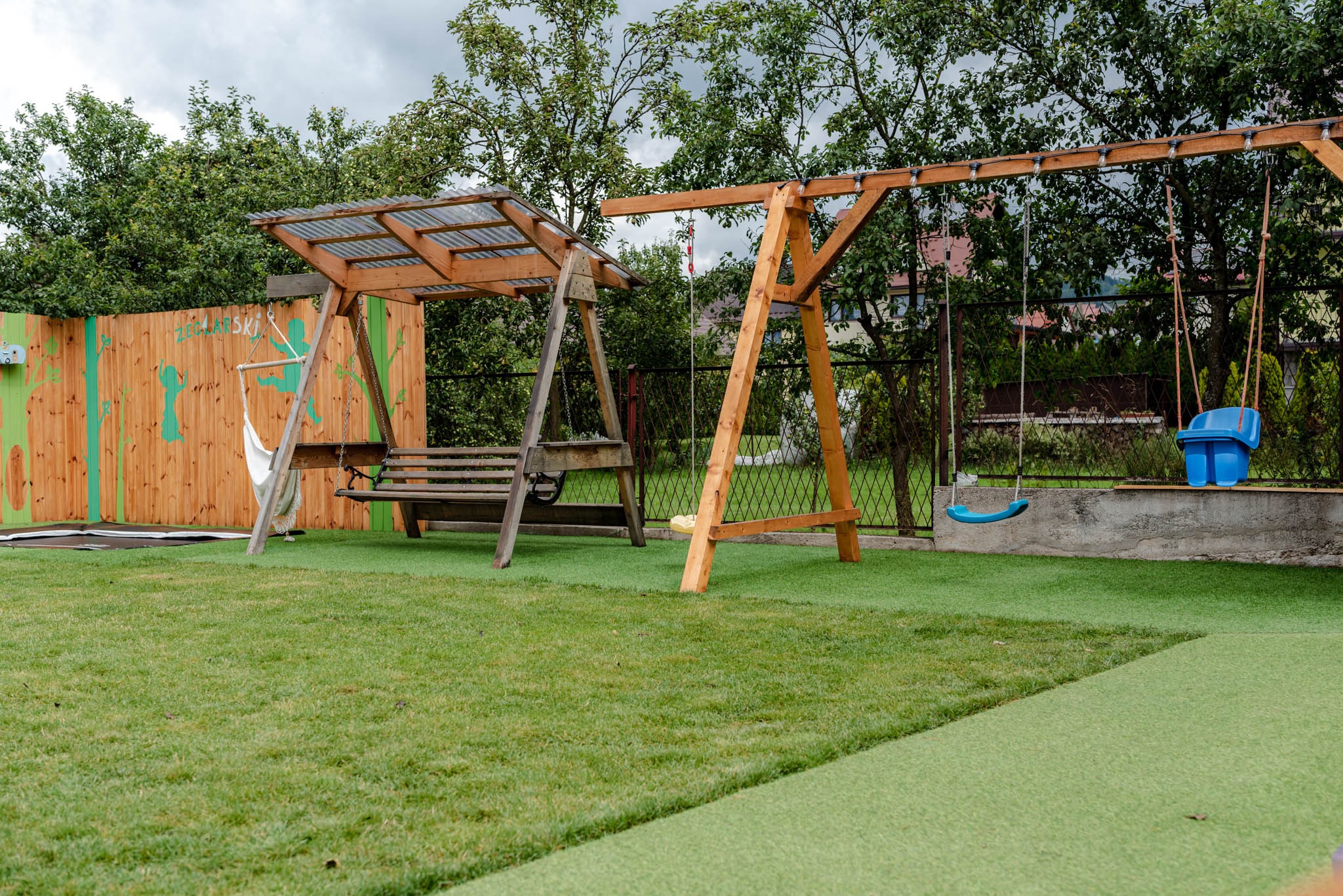 Drewniany plac zabaw z huśtawkami i hamakiem na zielonej trawie, ogrodzony drewnianym płotem z malunkami.