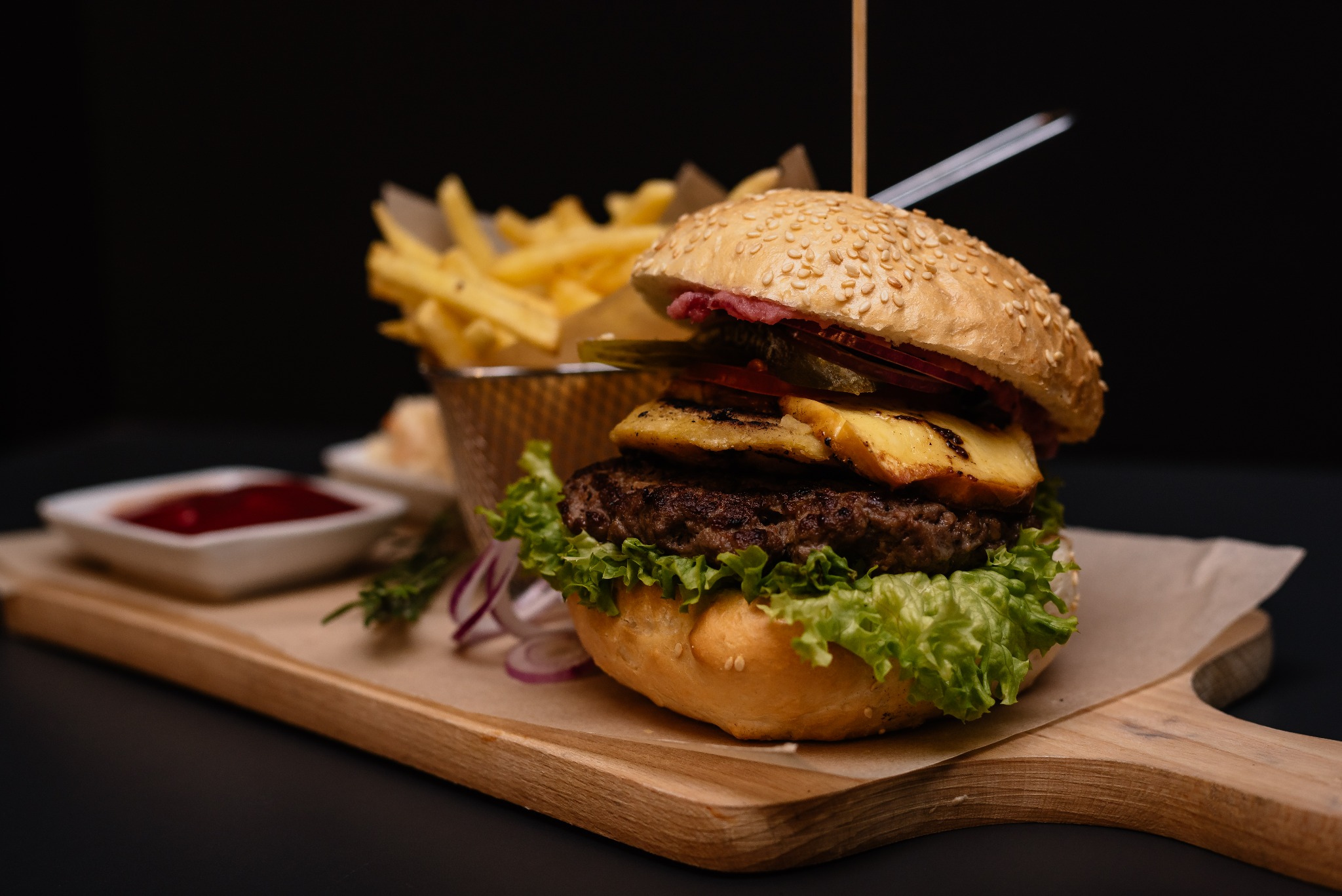 Apetycznie wyglądający burger z wołowiną, serem, sałatą i dodatkami, podany z frytkami w metalowym koszyczku i sosem na drewnianej desce na czarnym tle.