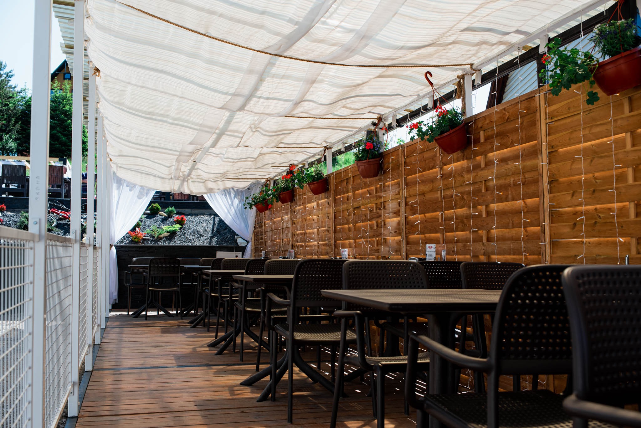 Zadaszony taras restauracji z drewnianą podłogą, czarnymi krzesłami i stołami, dekorowany kwiatami w wiszących donicach i delikatnymi lampkami.