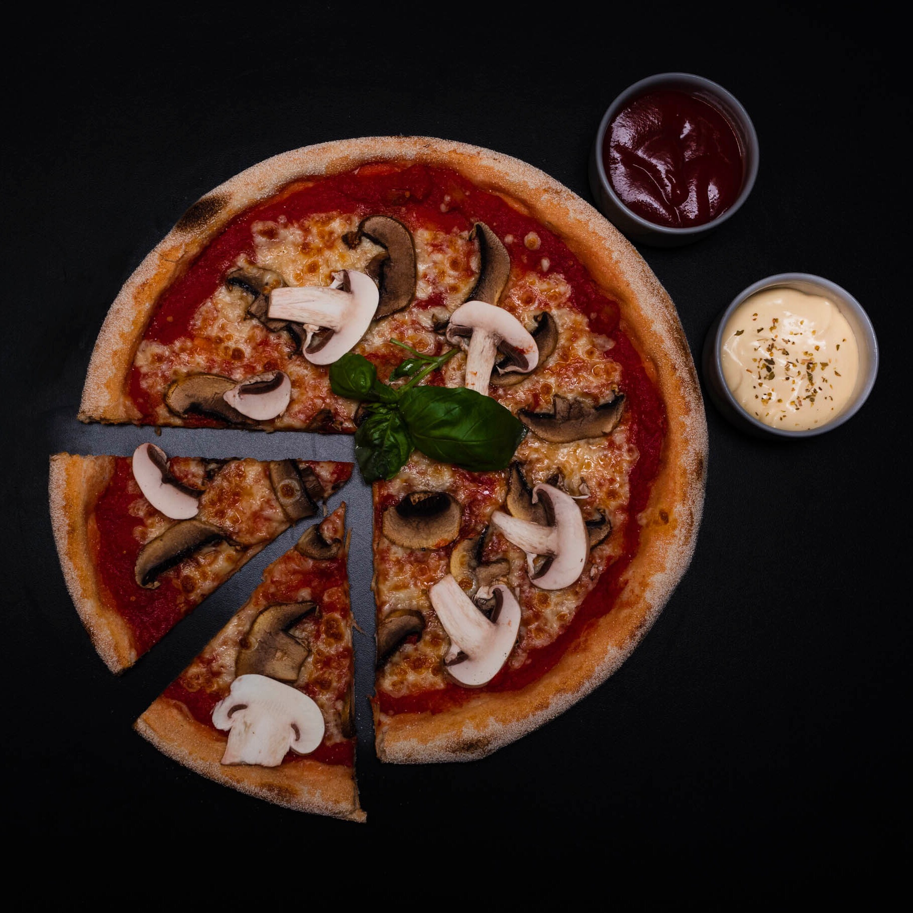 Pizza z pieczarkami, bazylią i dwoma sosami na czarnym tle, z wyciętymi dwoma kawałkami.